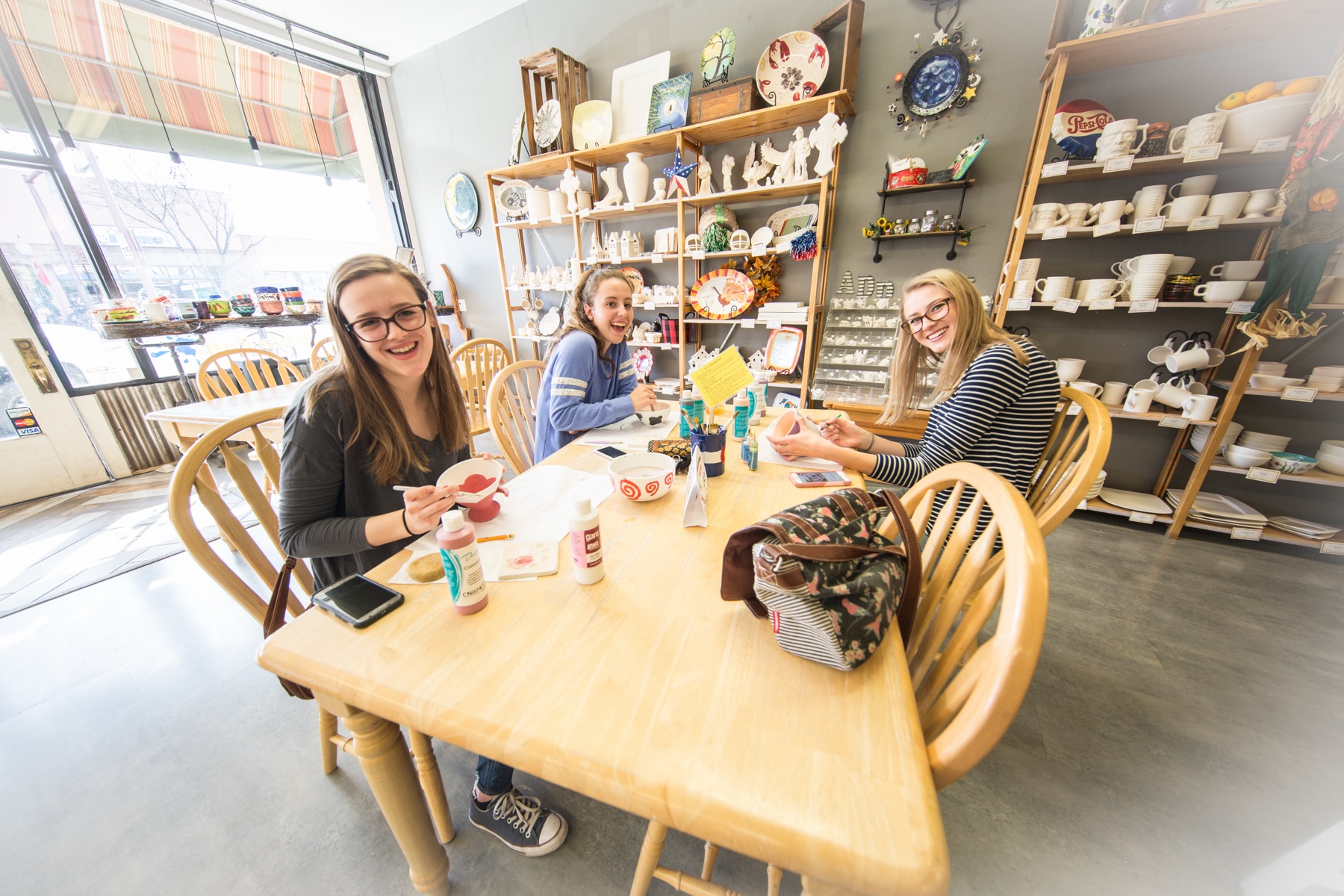 Guests painting pottery together at a long table inside Amazing Glaze studio, surrounded by shelves of ceramics